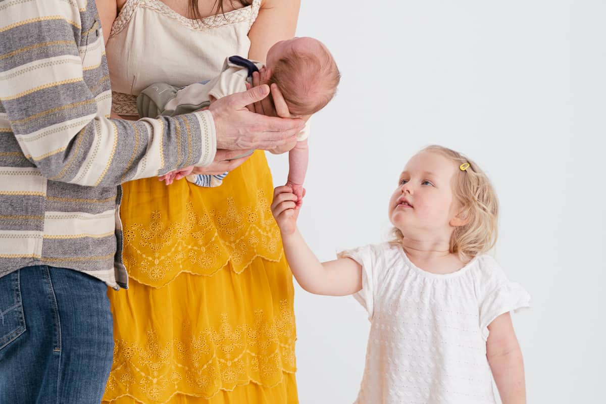 Toddler sister reaches for her newborn baby brother held by parents 