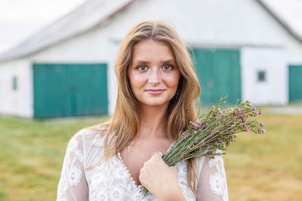 Senior girl poses in front of white barn with green doors, holding a spray of flowers