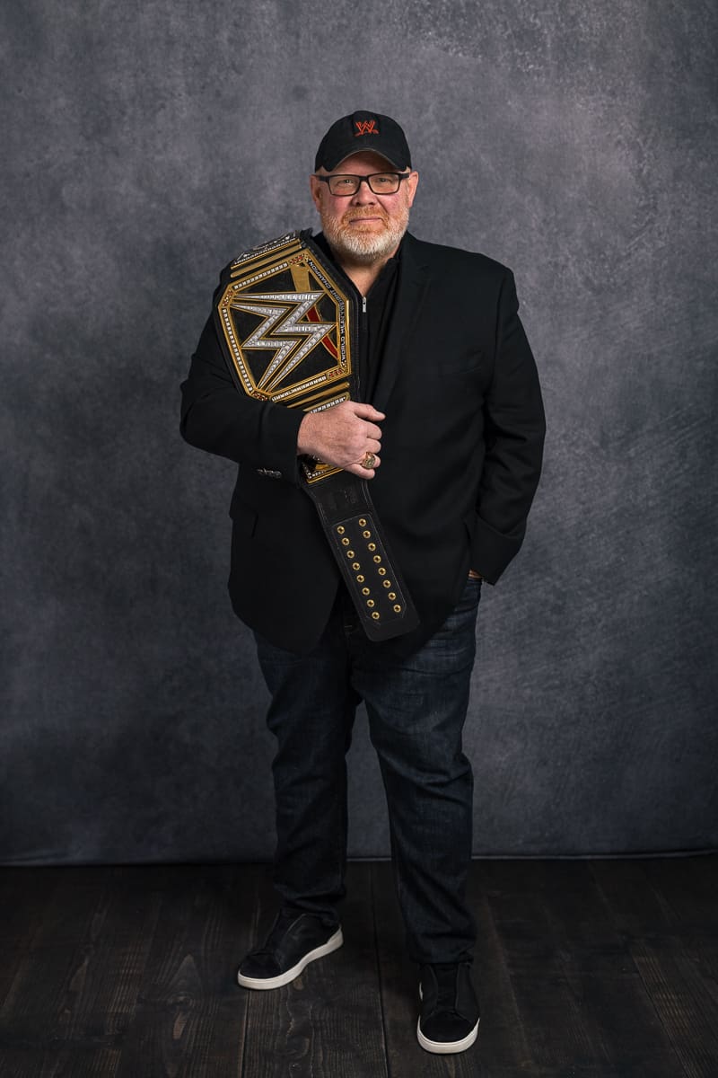 Man in baseball cap and black jacket and jeans holds championship wrestling belt