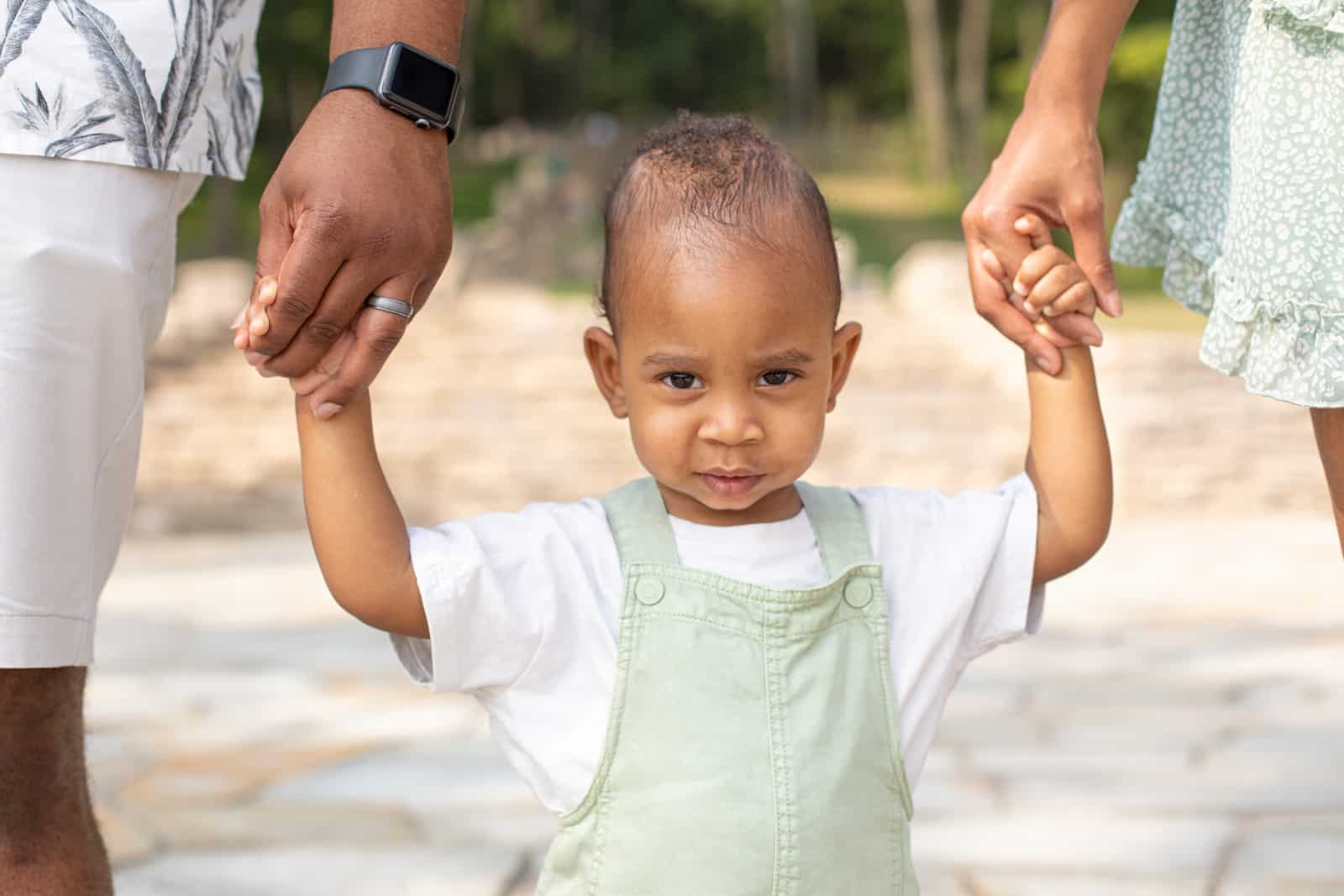 Black couple hold the hands of their one year old baby at the park.
