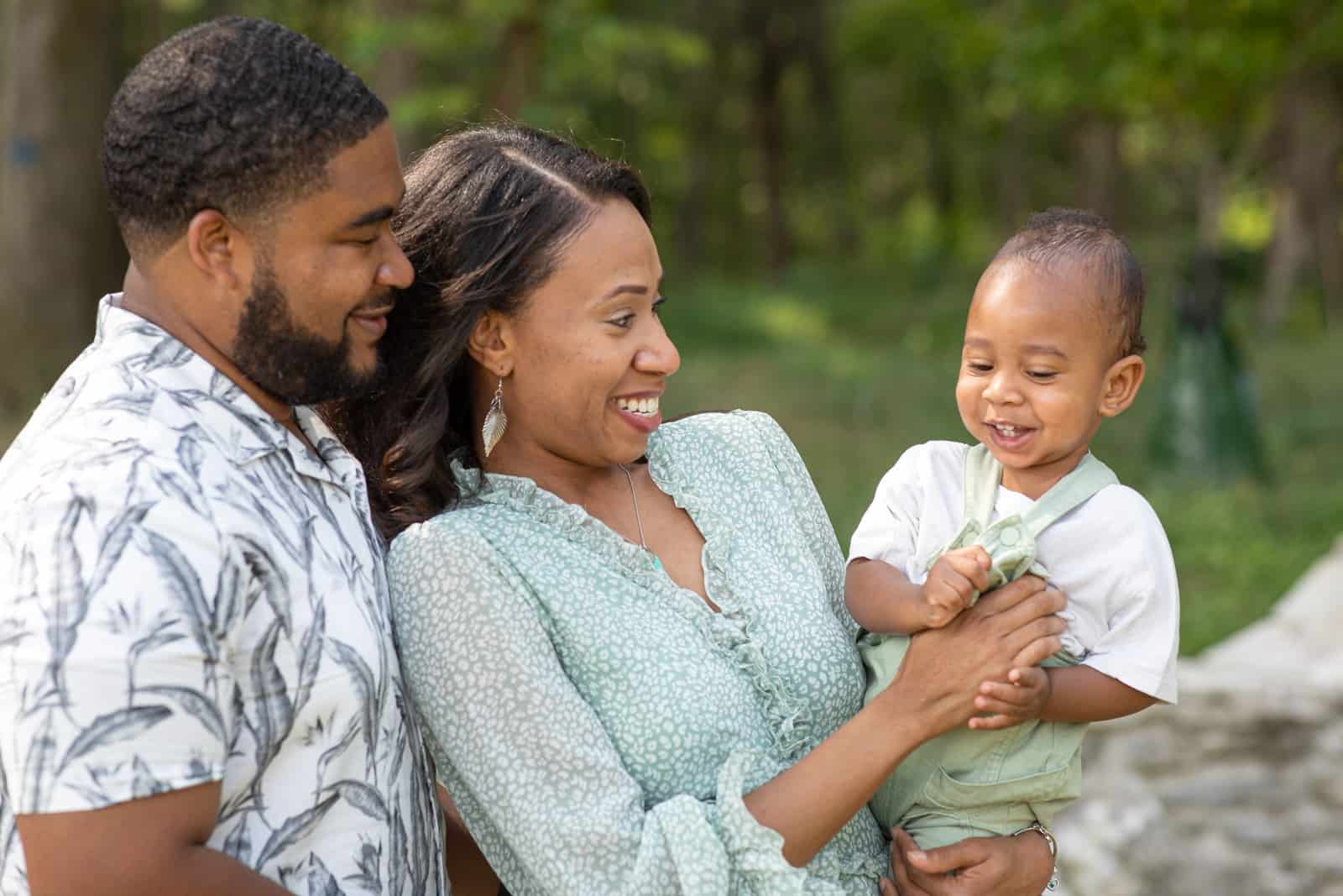 Black couple smiling and holding their one year old baby at the park.