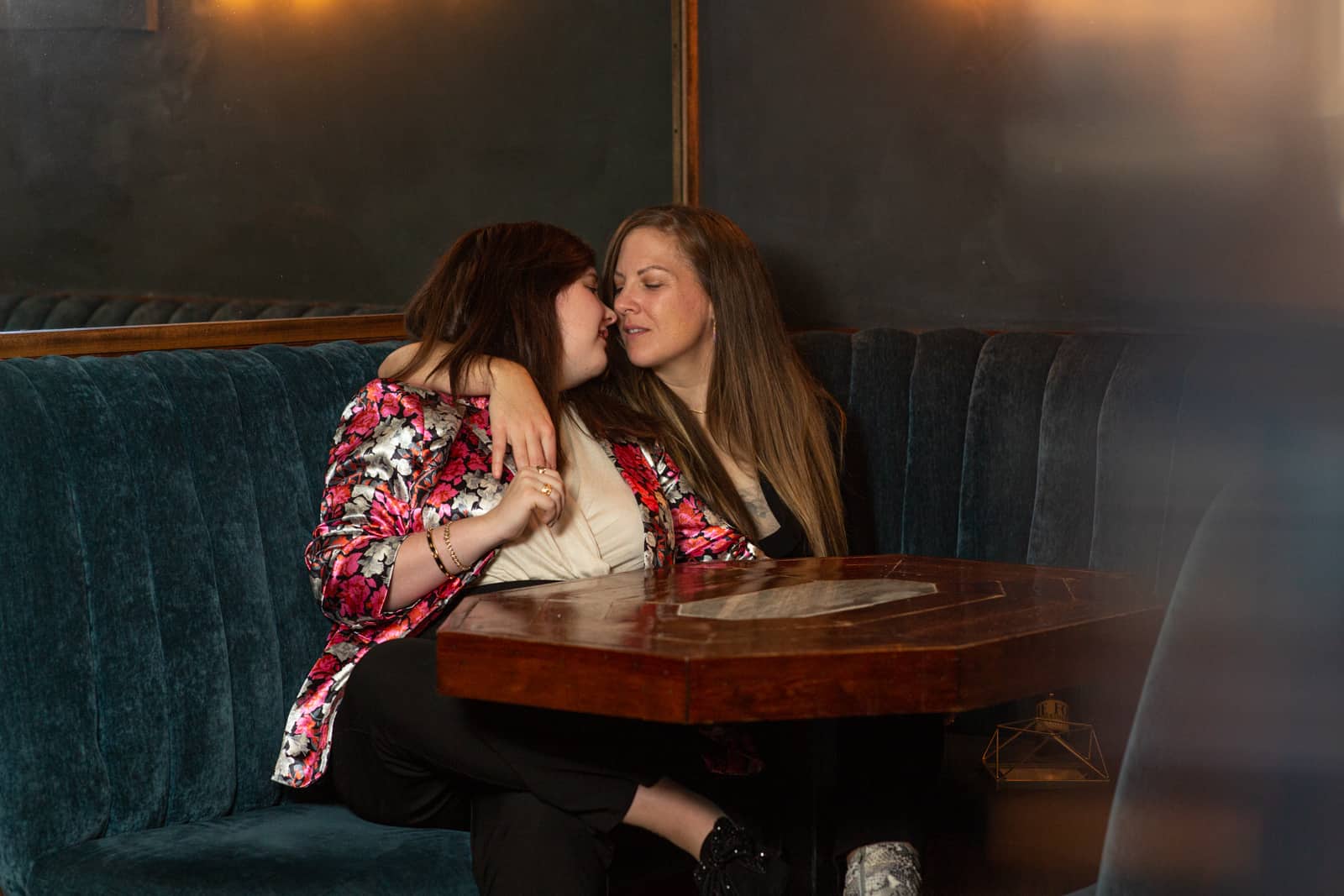 two women who are LGBTQ couple share a cozy corner booth inside a bar and hug