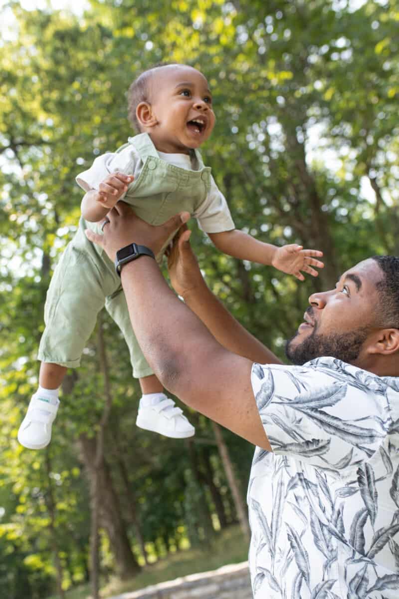 Black father holds his one year old son up in the air outside at the park.