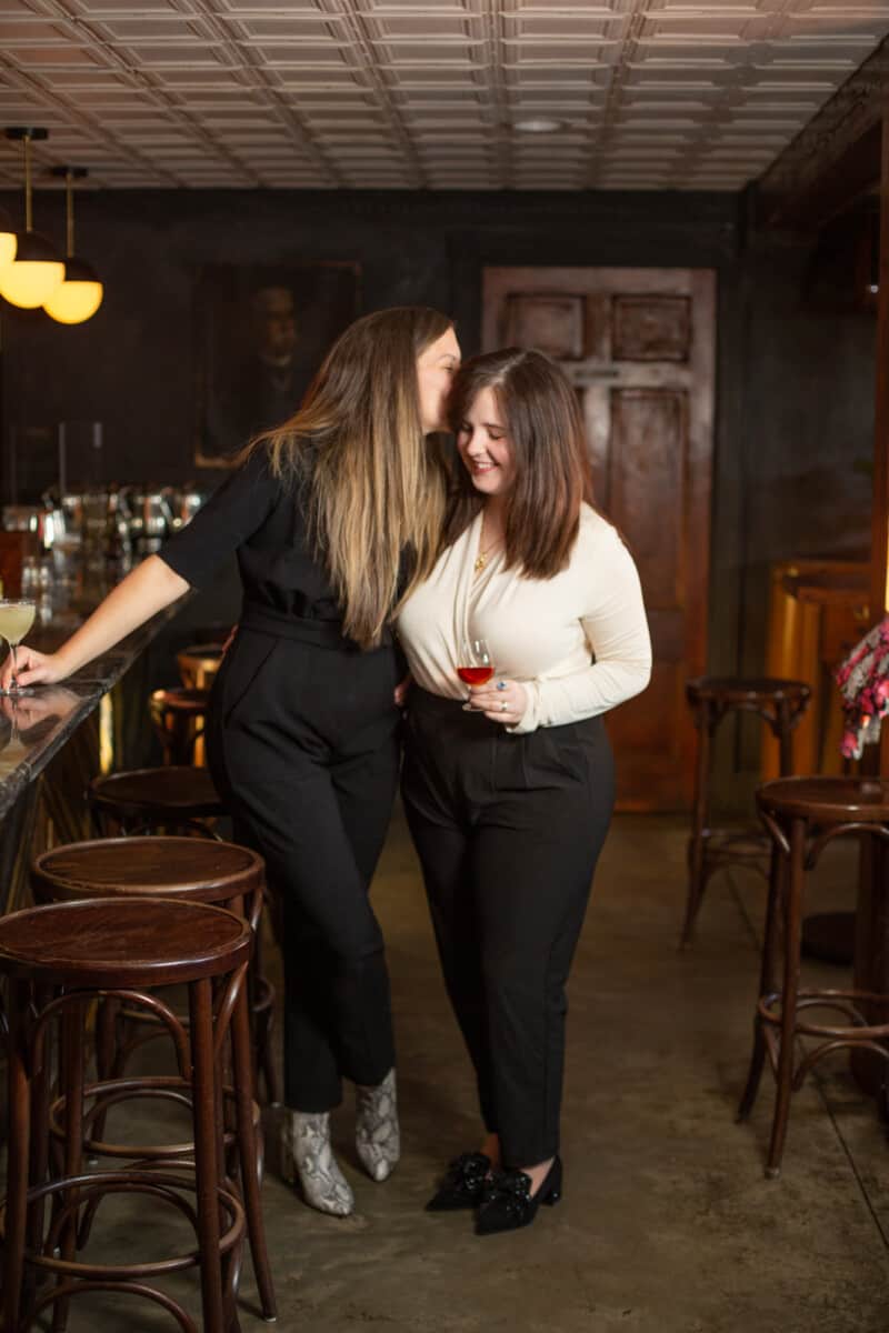 two women who are LGBTQ couple in dimly lit bar hold cocktails and whisper to each other
