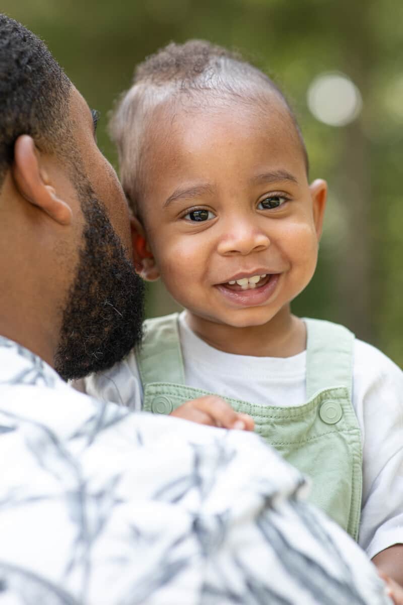 One year old black baby boy smiles as he is held by his father and looks over his shoulder.