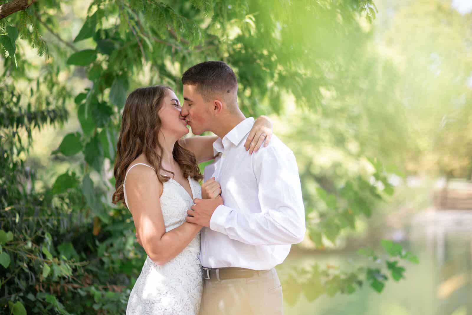 Elopement groom with white cotton shirt and bride with sleeveless lace dress kiss in Centennial Park surrounding by soft sun-filled foliage.