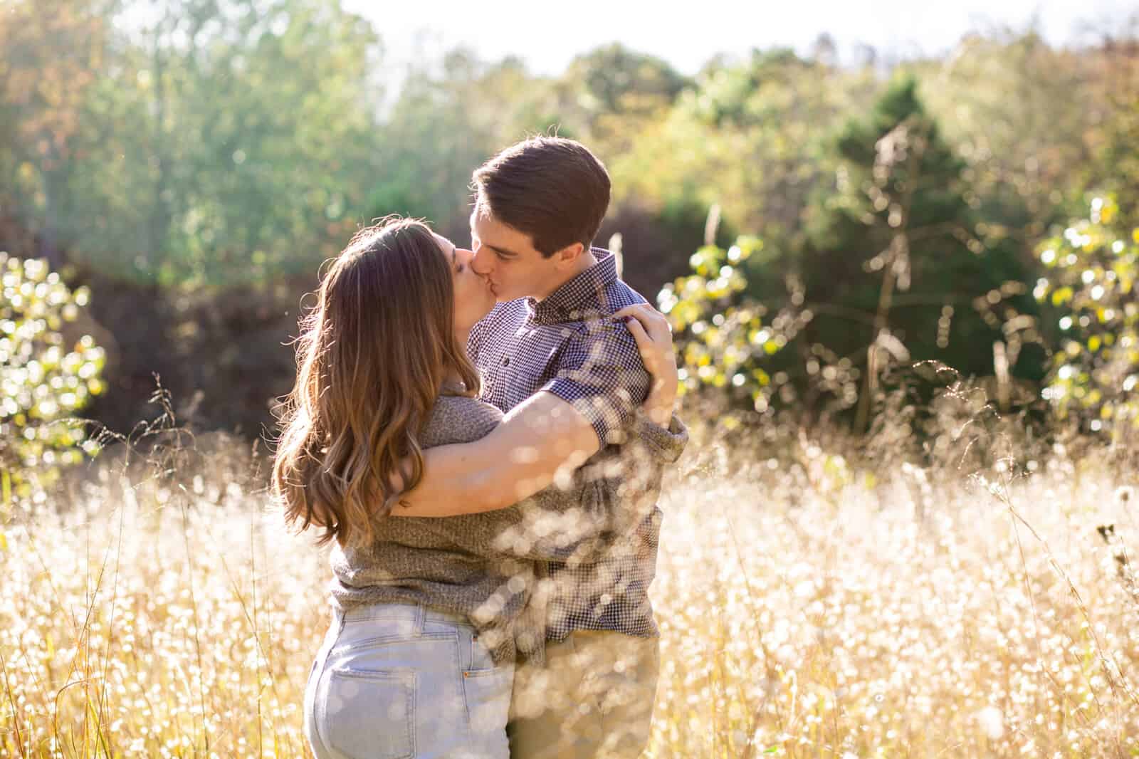 Young engaged girl with long brown hair kisses her fiance in a golden sunlit field.