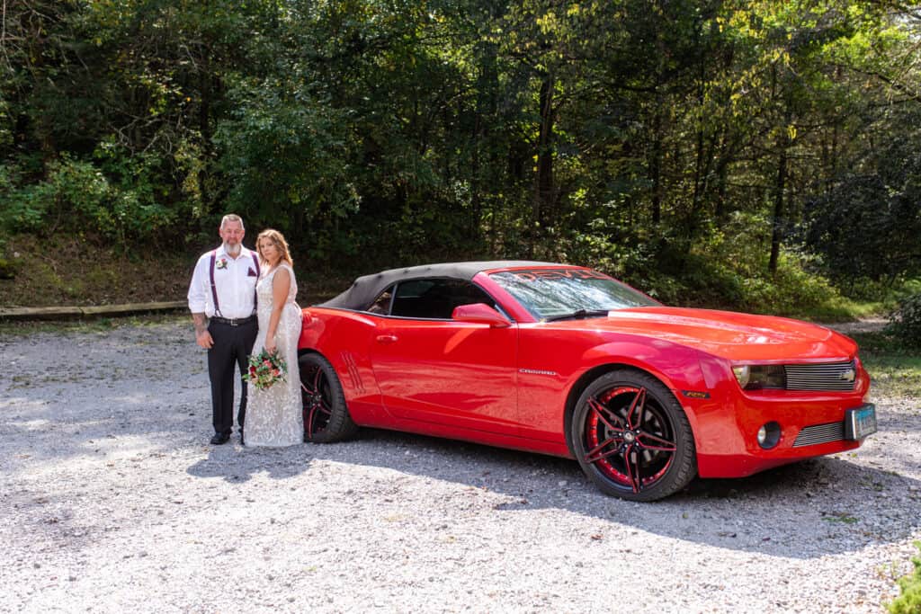 couple in wooded area pose with red Camero car