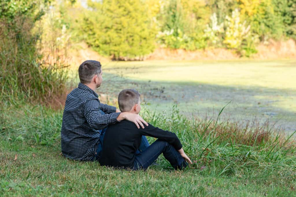 Father puts his arm around son as they sit near a pond