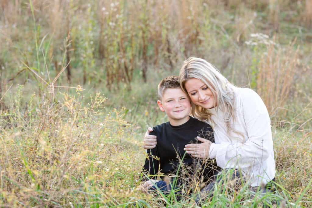 Blond mother with 10 year old son sitting in soft grassy field as she hugs him