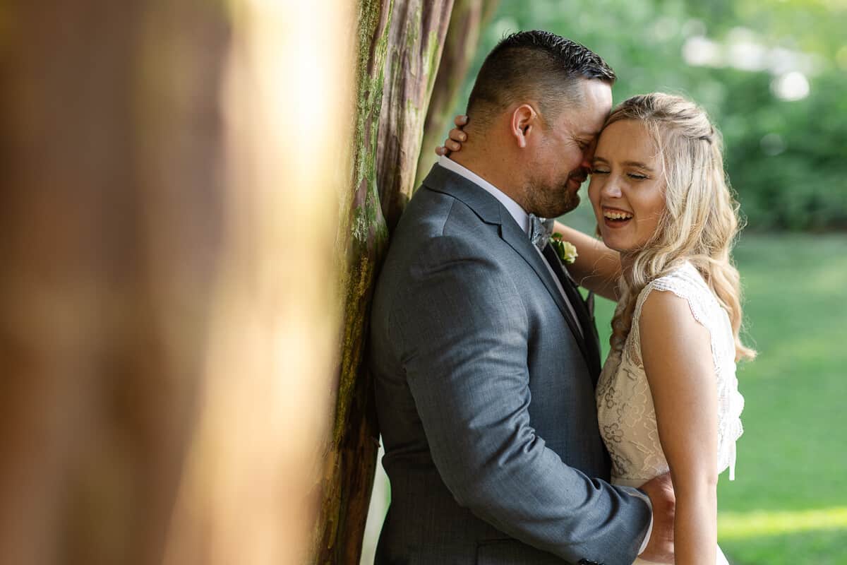 groom whispers in the ear of his bride as he leans against a tree