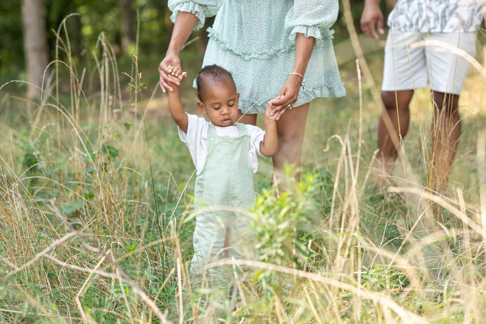 Black couple hold the hands of their one year old baby in tall grass at the park.