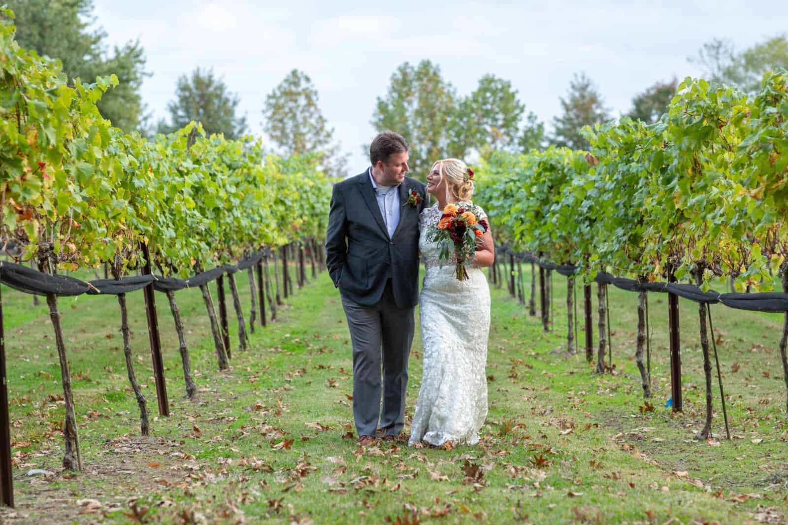 Bride and groom walk down middle of vineyard row with arms around each other.