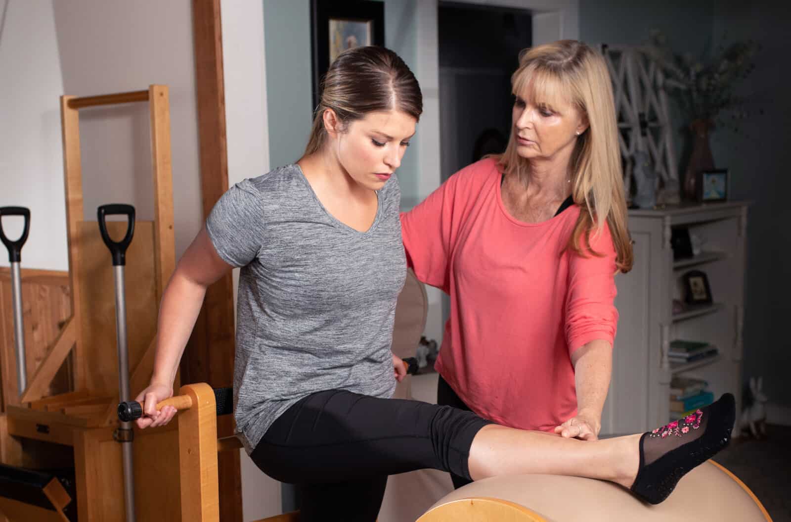Pilates teacher with bright coral shirt helps student stretch her leg over a piece of barrel equipment.