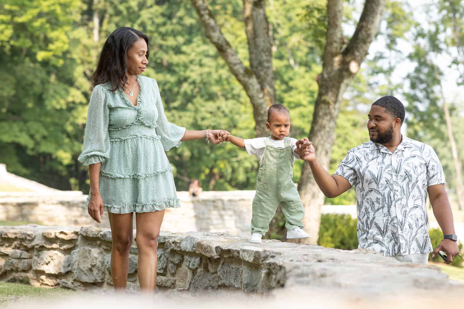 Black couple hold the hands of their one year old baby as he walks on a wall at the park.