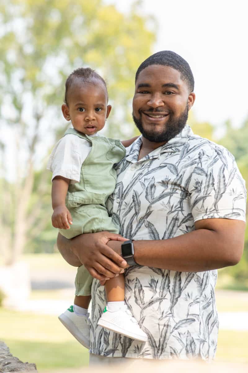 Black father holds his one year old son outside at the park.