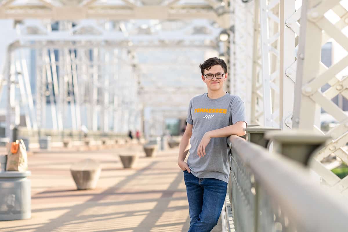 senior boy leans against railing while standing on bridge