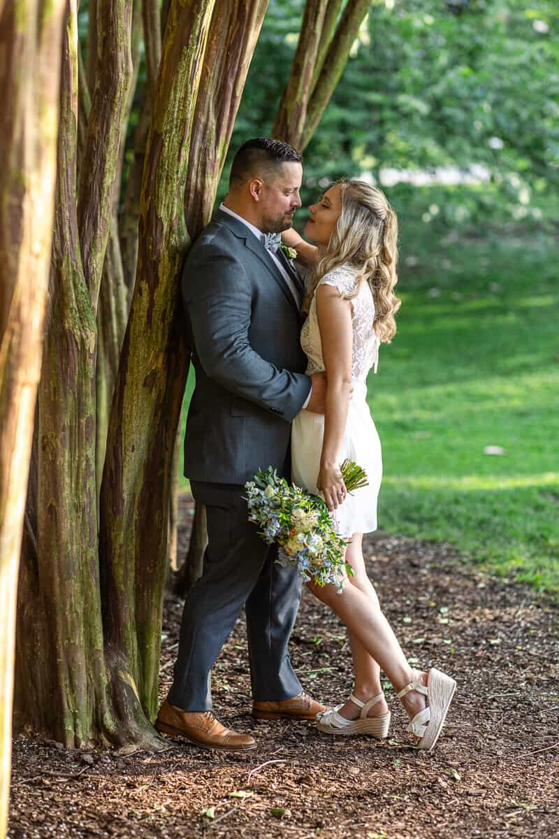 bride and groom hug as groom leans against a shaded tree