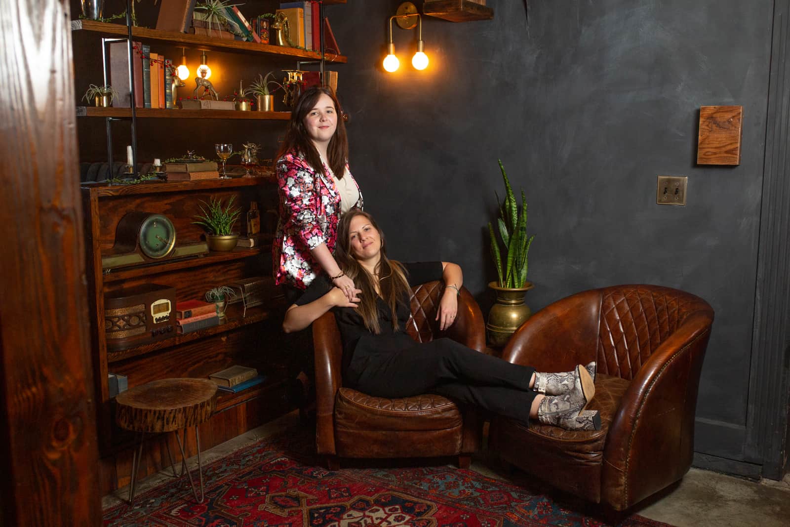 Two women posing inside a dark lit bar and sitting on lounge furniture