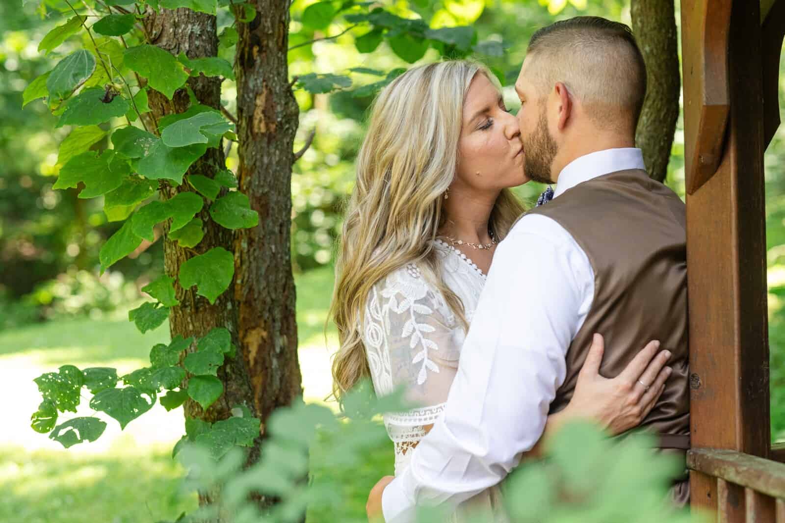 Bride and groom lean against gazebo and kiss beneath green leaves