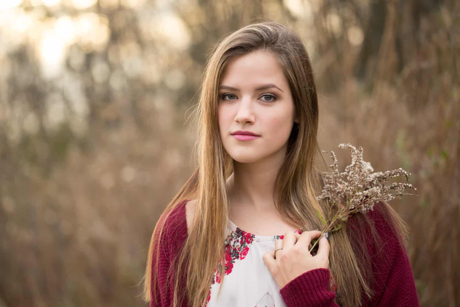 teen girl in burgandy sweater and flowered dress holds fuzzy flowered sprig in front of golden brown field.