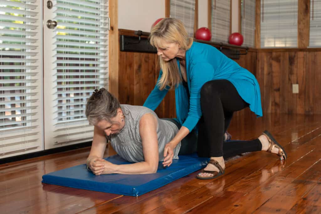 blond pilates instructor kneeling to assist student with mat pilates