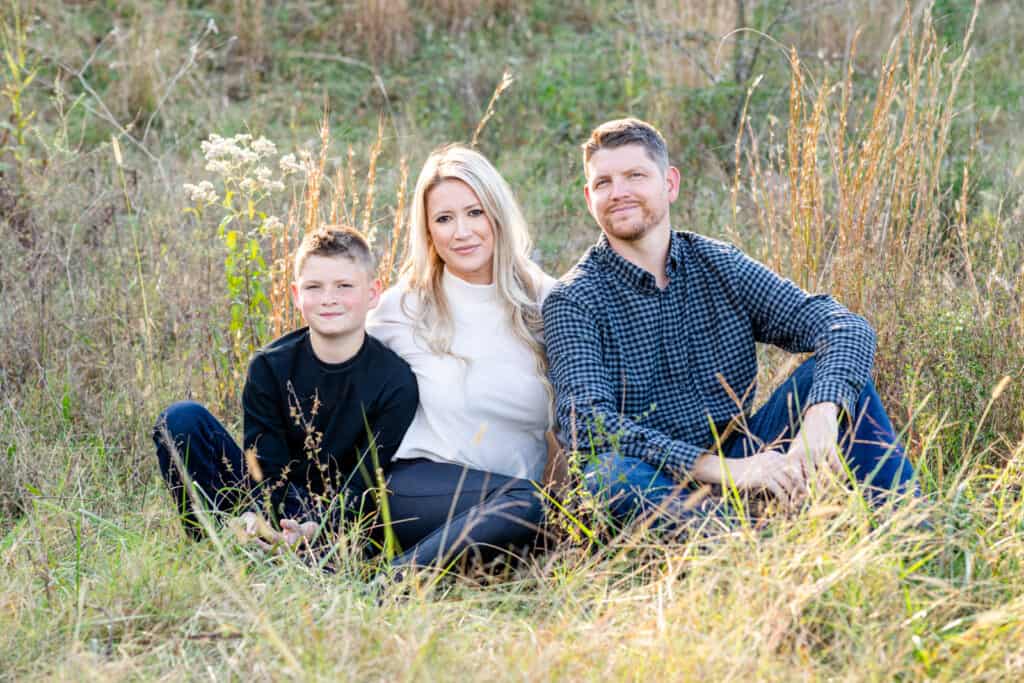Blond mom, dad and 10 year old son sit in light grassy field smiling at camera