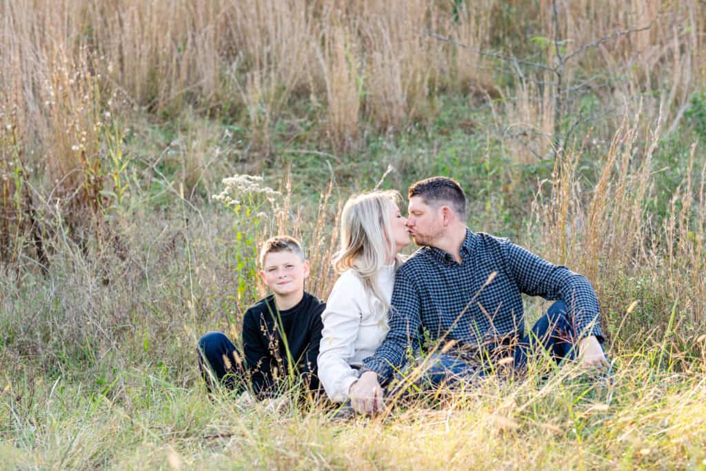 Family of three sits in field, blond mother kisses dad while 10 year old son looks at camera