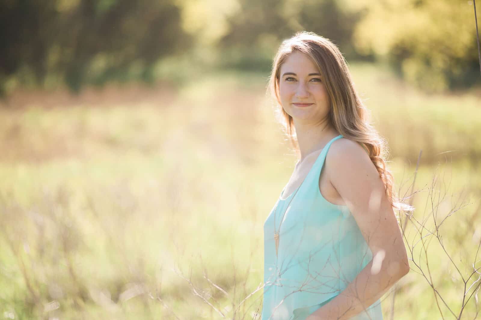 Teen girl with light brown long hair and aqua shirt stands in tall grass field for senior portrait on bright sunny day