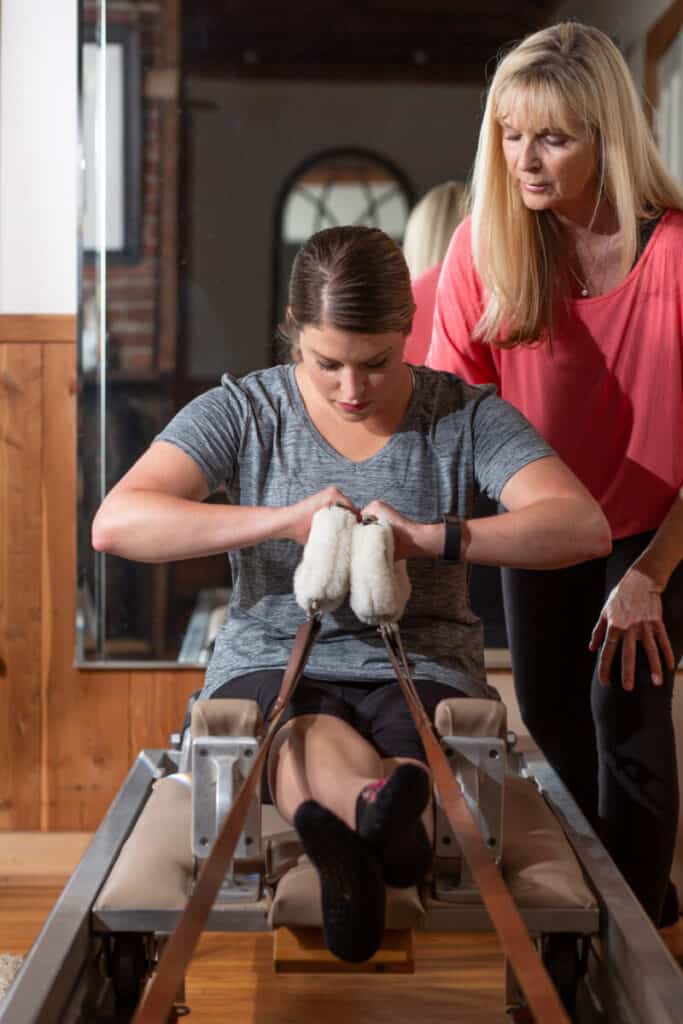 woman uses pilates equipment assisted by blond instructor