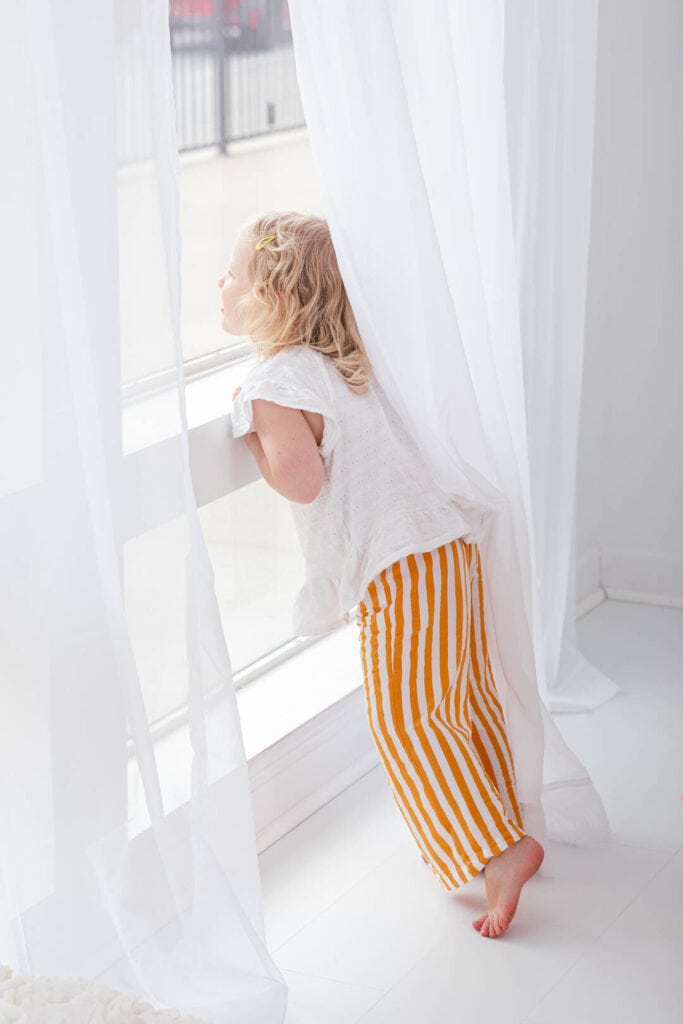 Toddler girl peers out window bathed in light