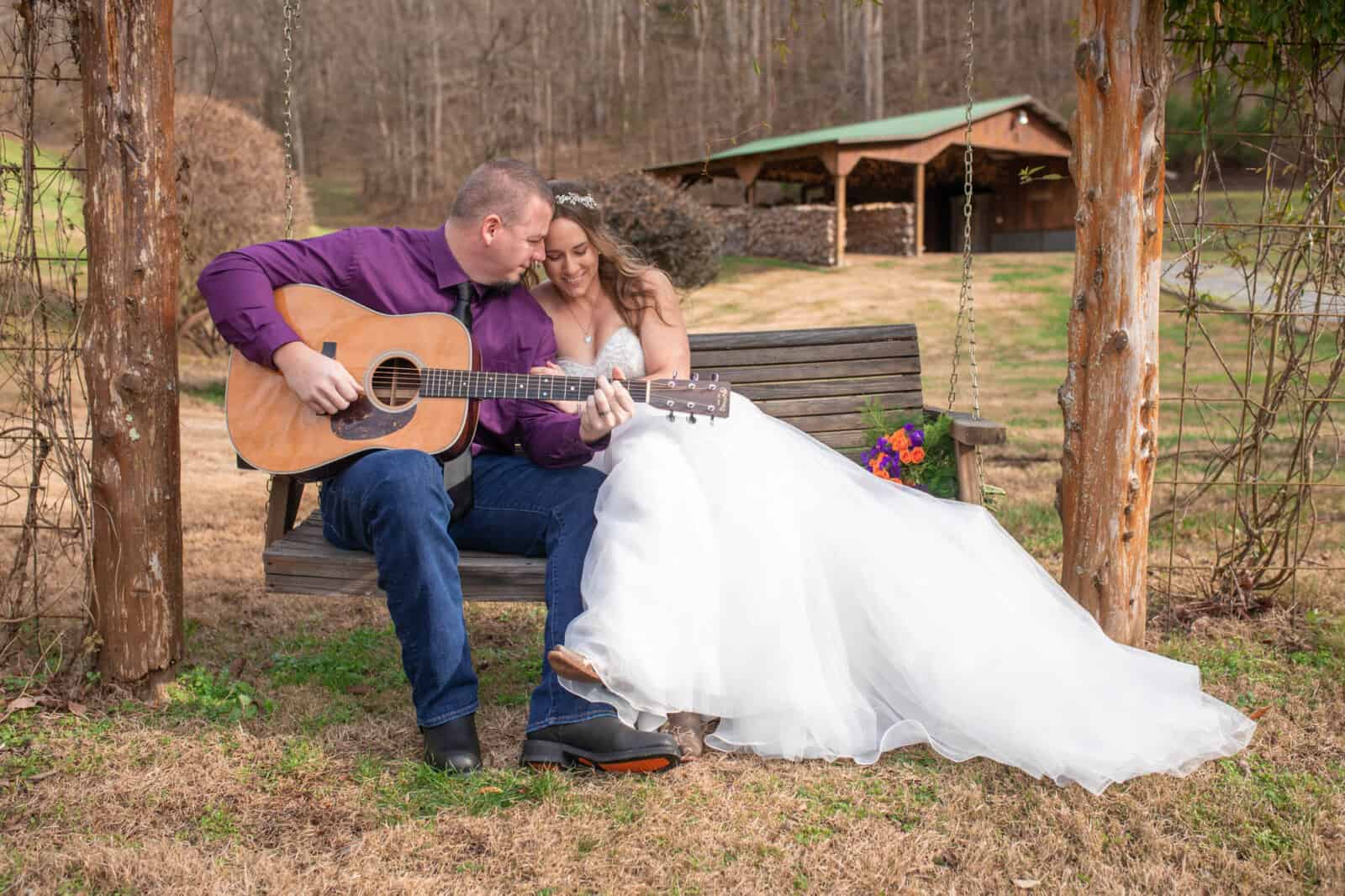 Elopement groom with purple shirt sings and plays acoustic guitar to bride in long flowing wedding gown sitting on swing near barn.
