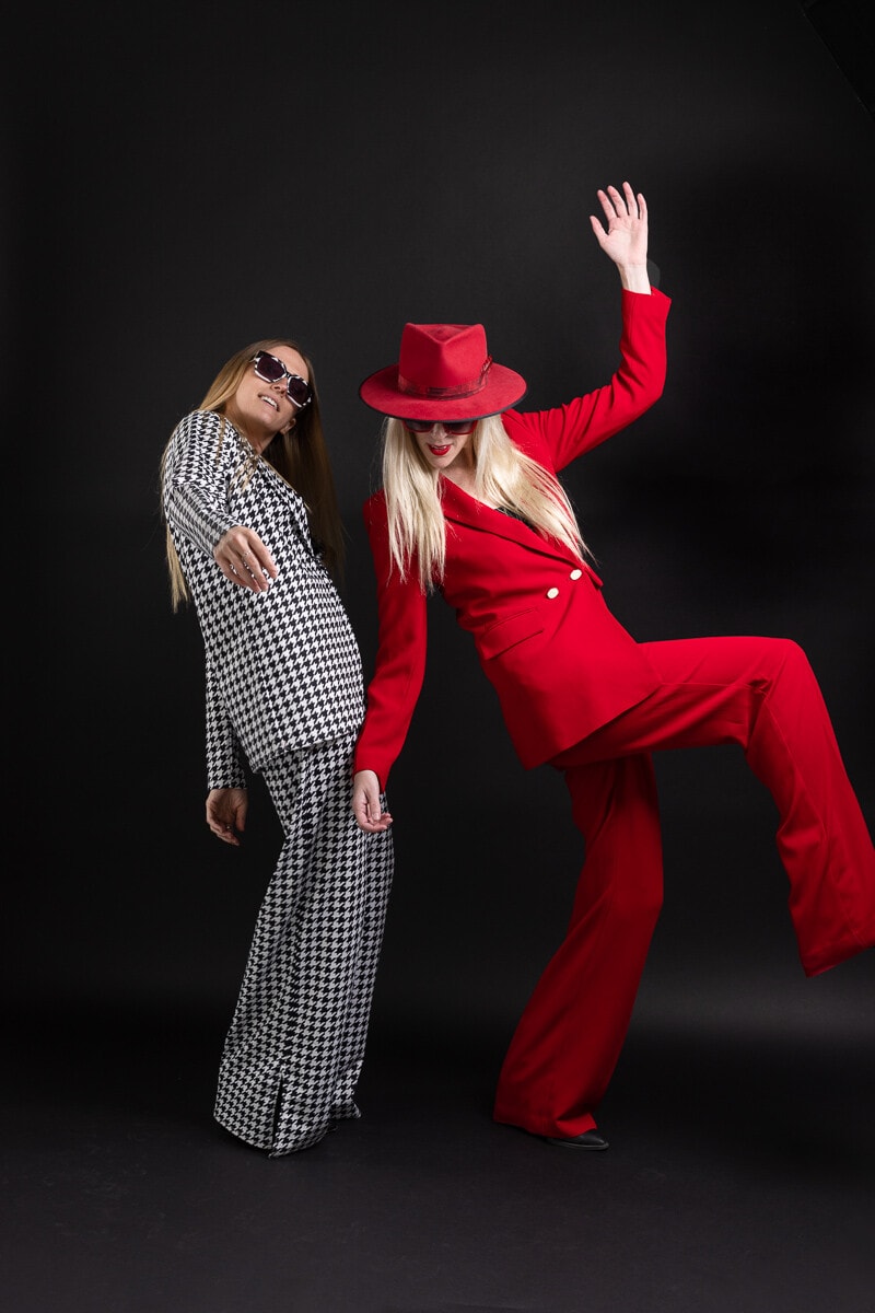 two women in fashion pantsuits dance against black background