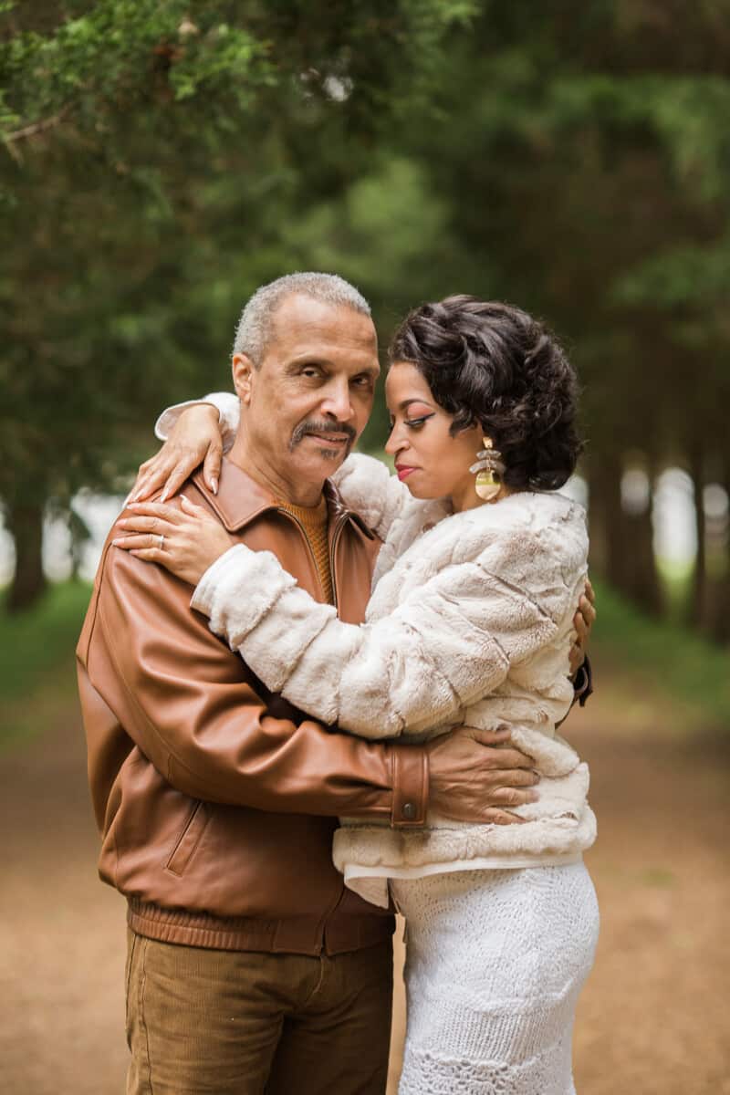 African American mature man in brown leather jacket embraces his fiance in white fur jacket in winter portrait session.