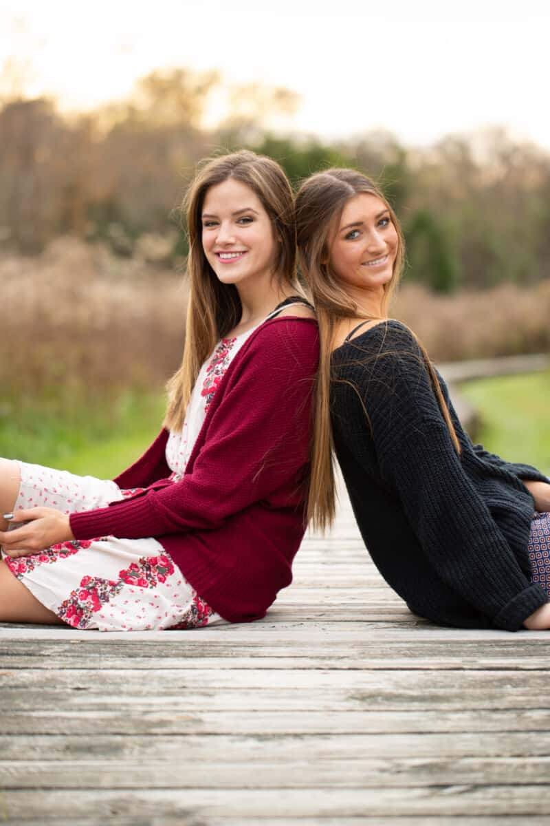 two teen girls with long hair sit back to back on wooden walkway smiling for senior portraits.